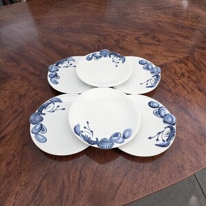 May include: A set of six white ceramic plates with a blue seashell and lobster design. The plates are arranged on a dark wooden table. The plates are a mix of round and oval shapes.