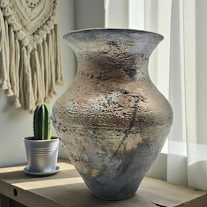 May include: A handmade ceramic vase with a textured surface, wide opening, and rounded shape. The vase displays a mix of earthy colours: brown, gold, and grey, with delicate dark patterns. A decorative item for home interiors.