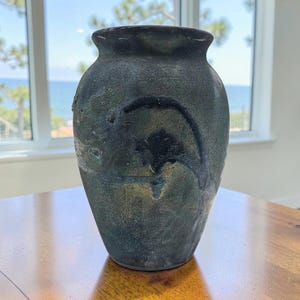 Pode incluir: Um vaso de cerâmica escuro e texturizado com um corpo largo e um pescoço estreito. O vaso tem uma aparência manchada com tons de verde, azul e preto. A superfície tem uma textura áspera e irregular, dando-lhe um aspecto rústico e artesanal. O vaso tem cerca de 30 cm de altura.