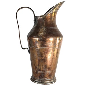 Op de afbeelding: Een vintage koperen kan met een gehamerde afwerking en een gebogen handvat. De kan heeft een verweerde, gepatineerde uitstraling en is in een rustieke, antieke stijl.