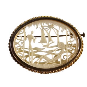 May include: An oval brooch carved from white material with a gold rope border. The brooch depicts a stylised scene of a person standing on a beach with sailboats in the background.