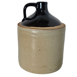 Puede incluir: Una jarra de barro vintage con la parte superior esmaltada en negro y un asa. La jarra es de color beige claro y tiene una textura rugosa.