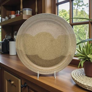 May include: A large, round ceramic plate with a speckled, earthy color palette. The plate features a textured surface and a wavy, layered design in shades of tan and brown. It is displayed on a white stand, suggesting a decorative or serving purpose.