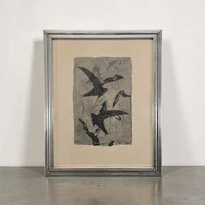 May include: A framed artwork featuring a dark, textured gray print of several birds in flight against a light beige background. The piece is enclosed in a metallic silver frame with a multi-tiered, stepped profile and a clean, rectangular border.