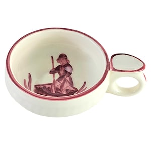 May include: A small white ceramic cup with a pink rim and handle. The inside of the cup features a hand-painted design of a person in a boat rowing with an oar.