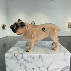 May include: A ceramic bulldog sculpture in a standing pose, with a tan body and black accents on the face and ears. A small, circular emblem is affixed to its side. The sculpture is displayed on a white marble block.