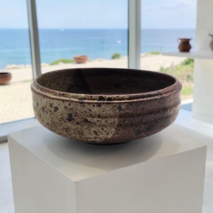 May include: A handcrafted ceramic bowl with a wide, shallow design. The bowl features a two-tone color scheme, with a light beige exterior speckled with dark spots and a dark brown interior. The rim has a subtle ridge.