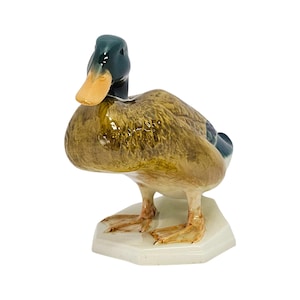 May include: A glossy ceramic mallard duck figurine featuring a dark green head, a light yellow beak, and a brown mottled body. The duck stands on a white, hexagonal base, with its head turned slightly to the left, showcasing a realistic posture.