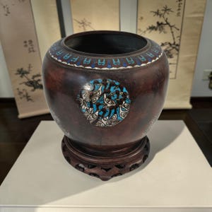 May include: A dark brown, antique-style vase with a wide opening and a decorative wooden base. The vase features a circular turquoise and white design on the side, and a blue and white pattern around the rim. The vase is likely made of metal.