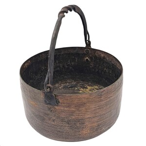 May include: A rustic, antique-style copper cauldron with a dark, twisted metal handle. The cauldron has a textured surface and a dark interior, suggesting age and use. The handle is attached with rivets. The overall color is a warm, aged copper.