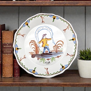 May include: White ceramic plate with a scalloped edge, featuring a hand-painted scene of a figure with a yellow jacket and blue trousers. The plate has a blue and green floral border, with the text "Vivre libre ou Mourir" and the number "1789".