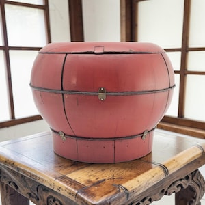 May include: A red, round, antique wooden container with a lid, metal bands, and brass clasps. The container is sitting on a carved wooden table. The container is approximately 18 inches in diameter.