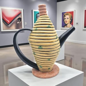 May include: A unique, abstract teapot sculpture with a light yellow, layered body and a black handle and spout. The base is a reddish-brown color, and the top features a stack of pink rings. The artwork is displayed in a gallery setting.