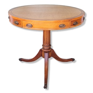 May include: A round wooden table with a leather top and three legs. The table has a drawer with brass hardware.