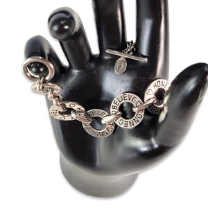 Pode incluir: Pulseira de prata com pingentes redondos. Os pingentes têm palavras como "Acreditar", "Conectar", "Apreciar", "Esperar" e "Respeitar".