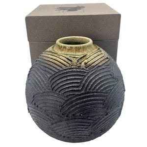 May include: A handcrafted, spherical ceramic vase with a textured, dark gray exterior and a light green interior. The vase is presented with a matching box, suggesting it's a gift item. The design features a unique, carved pattern.