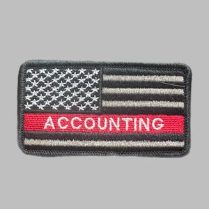 Peut inclure: Un écusson rectangulaire avec un motif inspiré du drapeau américain. Il présente des étoiles blanches sur fond noir, des rayures noires et grises, et une bande rouge avec le mot "ACCOUNTING" en lettres blanches.