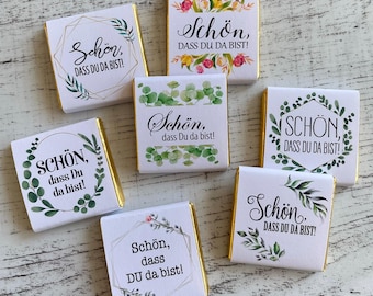 Puede incluir: Un conjunto de siete pequeñas barras de chocolate envueltas en papel de aluminio dorado con envoltorios de papel blanco con un diseño floral y el texto "Schön, dass du da bist!" en alemán, que se traduce como "¡Es agradable que estés aquí!"