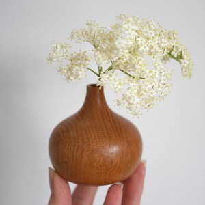 May include: A small, handcrafted wooden vase with a bulbous base and narrow neck, holding a sprig of delicate white flowers. The vase is a warm, natural brown colour, and the flowers provide a bright contrast. The vase is approximately 10 cm tall.