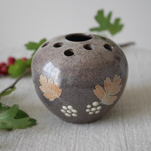 May include: A spherical, speckled grey ceramic vase with multiple holes on top for flower stems. The vase is decorated with leaf and berry designs in tan and white. A sprig of red berries and green leaves is in the background.