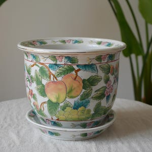 May include: A white ceramic planter with a matching saucer, featuring a hand-painted design of colourful apples, green leaves, and clusters of grapes. The rim is decorated with a repeating floral pattern and a thin gold-coloured trim detail.
