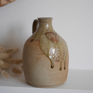 May include: A beige ceramic jug with a brown glaze and a brown crest on the front. The glaze has dripped down the side of the jug.