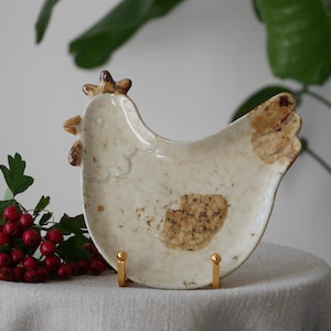 May include: A cream-coloured ceramic chicken-shaped plate with brown markings, supported by two small gold stands. A sprig of red berries and green leaves are to the left of the plate. The plate is likely for serving food.