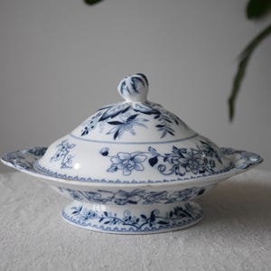 May include: A white porcelain serving dish with a lid, decorated with blue floral patterns. The dish has a round base and a matching lid with a small knob. The design features detailed blue flowers and foliage, creating an elegant aesthetic.