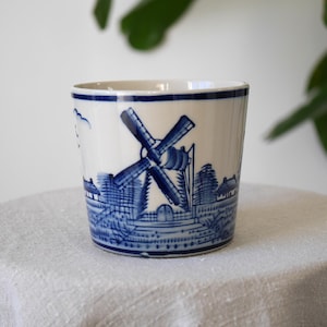 May include: White ceramic cup with a blue windmill and landscape design. The cup has a blue rim and a detailed illustration of a windmill with surrounding buildings. The design is reminiscent of Delftware pottery.