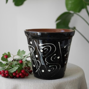 May include: A black ceramic plant pot with white abstract designs. The pot has a brown interior and sits on a neutral-coloured surface. A sprig of red berries and green leaves rests beside the pot, with a leafy plant in the background.