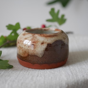 May include: A small, rounded ceramic pot with a wide opening. The pot has a layered design with a reddish-brown base, a dark brown middle, and a creamy, speckled top. The pot is likely handmade and suitable for small plants or decor.