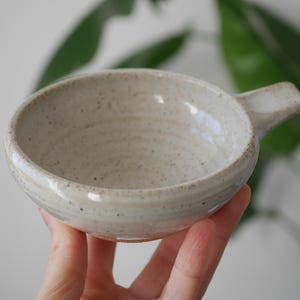 May include: A small, handmade ceramic bowl with a handle. The bowl is off-white with speckled detailing and a glossy finish. The handle is rectangular and extends from the side of the bowl. The bowl is held in a person's hand.