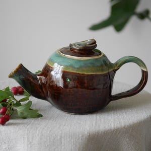May include: A handmade ceramic teapot with a glossy finish. The teapot has a brown base, with a green and teal rim and handle. The lid has a small, decorative knob. A sprig of red berries and green leaves is placed nearby.