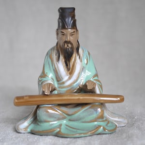 May include: A ceramic figurine of a man in a green and brown robe playing a long, brown musical instrument. He is wearing a black hat and has a long beard.