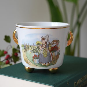 May include: A white porcelain bowl with two gold-toned handles and three gold-toned feet. The exterior features a colourful illustration of a person in traditional Dutch attire holding yellow flowers, surrounded by tulip buckets and a windmill.