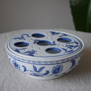 May include: A white and blue ceramic flower frog with a lid. The lid has several holes for arranging flowers. The bowl and lid are decorated with blue floral patterns. The flower frog is sitting on a white surface.