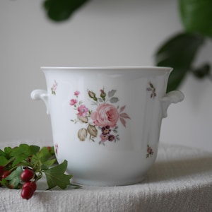 May include: White ceramic planter with two small handles and a floral design featuring pink roses and green leaves. The planter is decorated with a delicate pattern of pink and purple flowers. A sprig of red berries and green leaves rests in front of the planter.