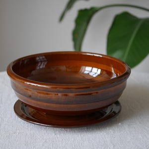 May include: A round, glazed ceramic bowl in a deep, glossy brown finish with a matching circular saucer base. The bowl features a wide, open top and a smooth, reflective surface, resting securely on the flat, coordinating brown ceramic plate.