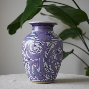 May include: A ceramic vase with a white base and a purple, abstract swirl design. The vase has a wide body, a narrow neck, and a flared rim. The vase is approximately 25 cm tall.