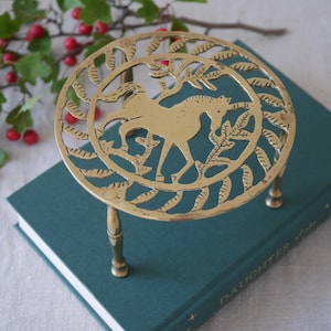 May include: A circular brass trivet featuring an intricate cutout design of a unicorn surrounded by stylised leaves. The trivet is supported by three slender metal legs and has a polished gold-toned finish with visible metallic texture.