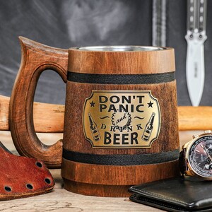 May include: A wooden beer stein with a black band and a gold metal plaque that reads "Don't Panic and Drink Beer". The stein is sitting on a wooden table with a leather wallet, a watch, and an axe.