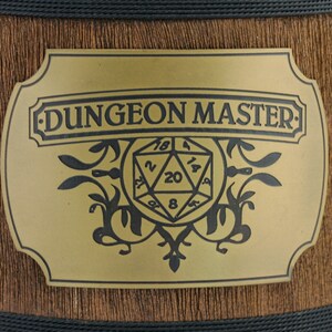 May include: A gold-colored metal plaque with a black inscription that reads "DUNGEON MASTER" and a black and white illustration of a twenty-sided die.