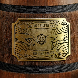 May include: A gold-coloured metal plaque with a black background and a design of two hands reaching towards a 20-sided die. The text on the plaque reads "The Dice Giveth And The Dice Taketh All."