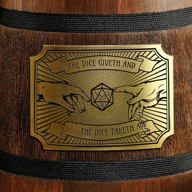 May include: A gold-coloured metal plaque with a black background and a design of two hands reaching towards a 20-sided die. The text on the plaque reads "The Dice Giveth And The Dice Taketh All."