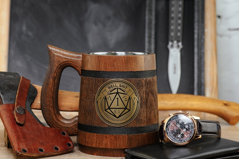 May include: A wooden beer stein with a stainless steel interior. The stein has a black band around the top and a gold metal emblem with a dice and the text "Well, Shit" on the front.