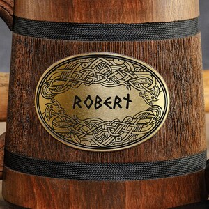 Pode incluir: Um copo de cerveja de madeira com uma faixa preta ao redor da parte superior e inferior. O copo tem uma placa de lat&atilde;o com um design de n&oacute; celta e o nome "ROBERT" gravado nela.
