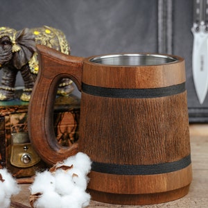 May include: A wooden beer stein with a stainless steel liner and a black band around the middle. The stein has a wooden handle and is sitting on a wooden surface.