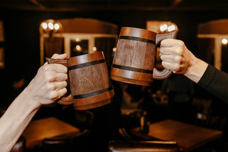 Pode incluir: Duas pessoas brindando com canecas de cerveja de madeira. As canecas s&atilde;o marrons com uma faixa marrom escura no meio.