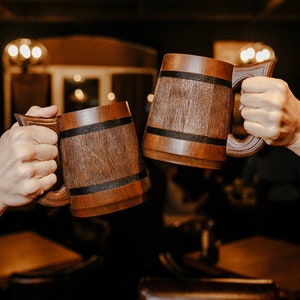 May include: Two people clinking wooden beer mugs together. The mugs are brown with a dark brown band around the middle.