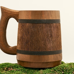May include: A wooden beer stein with a dark brown finish and a black band around the top. The stein has a large handle and is sitting on a bed of green moss.